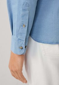 Persona con una camisa de manga larga azul claro con puños abotonados y pantalones blancos con costuras del bolsillo trasero visibles.