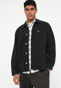 Giacca in denim nero con colletto, bottoni frontali, due tasche grandi e un piccolo accento di logo verde. Indossata sopra una camicia a quadri.