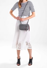 Šedá kožená crossbody taška se zipovým uzávěrem, hladkým povrchem a dlouhým nastavitelným popruhem. Kombinována s bílou skládací sukní a pruhovaným topem.