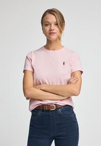 Rosa T-Shirt mit kurzen Ärmeln aus Baumwolle, mit einem kleinen schwarzen Logo auf der linken Seite. Kombiniert mit dunkelblauen High-Waist-Jeans und einem braunen Gürtel.