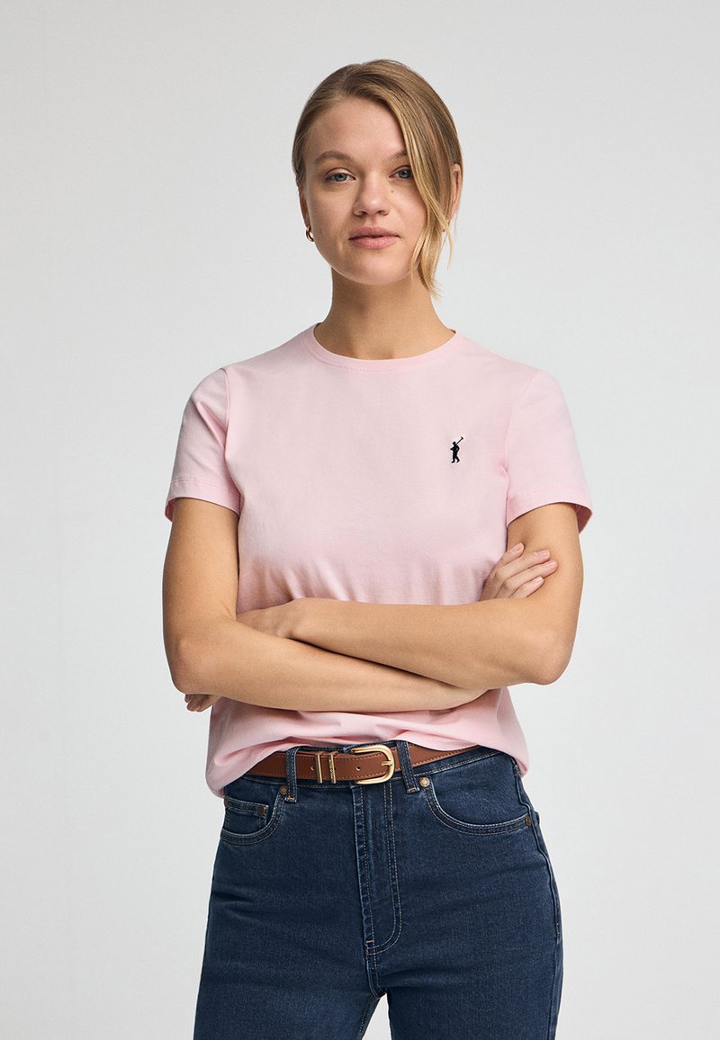 Rosa T-Shirt mit kurzen Ärmeln aus Baumwolle, mit einem kleinen schwarzen Logo auf der linken Seite. Kombiniert mit dunkelblauen High-Waist-Jeans und einem braunen Gürtel.
