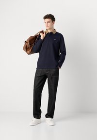 Polo a maniche lunghe blu navy con colletto in velluto a coste marrone, abbinato a pantaloni in denim scuro e sneakers bianche. Borsa in pelle marrone tenuta nella mano destra.