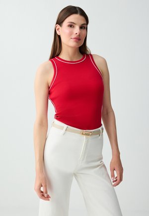 Donna con top rosso senza maniche con piping bianco e pantaloni bianchi a vita alta con cintura beige, in piedi davanti a uno sfondo uniforme.