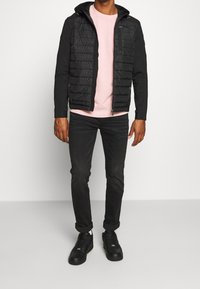 Veste noire matelassée avec une capuche, présentant un design matelassé. En dessous, un sweat-shirt rose est porté avec un jean noir et des baskets noires.