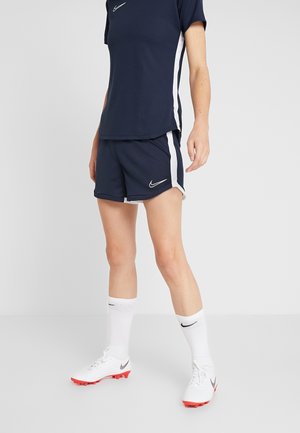 Person iført navyblå Nike sports-T-shirt og shorts med hvide striber, hvide sokker og hvide fodboldsko med røde såler.