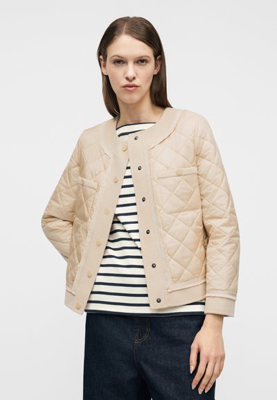Γυναίκα που φοράει μπεζ μπουφάν με καπιτονέ ύφασμα πάνω από μια λευκή και μαύρη ριγέ μπλούζα και σκούρο μπλε τζιν, στέκεται με το ένα χέρι στην τσέπη.
