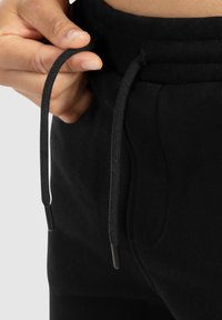 Schwarze Jogginghosen mit weicher Textur, ausgestattet mit einem Kordelzugbund und schwarzen Kunststoffenden. Nahaufnahme des Taillenbundes.