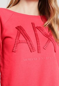 Sudadera roja con cuello de canalé y logotipo texturizado. Presenta "Armani Exchange" impreso debajo del logotipo con un acabado suave.