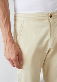 Lys beige bukser laget av myk stoff med en glatt tekstur, med knappelukking og skrå lommepanel.