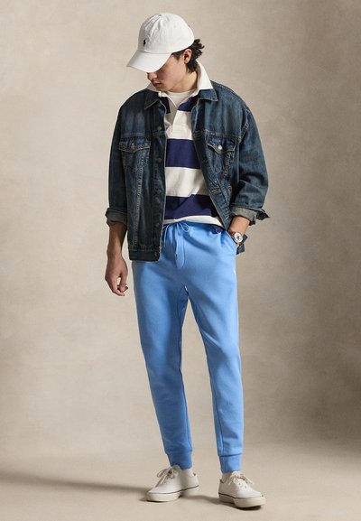 Mężczyzna w białej czapce, niebiesko-białej koszuli w paski, ciemnej kurtce dżinsowej, jasnoniebieskich spodniach dresowych, białych trampkach, stojący z jedną ręką w kieszeni.