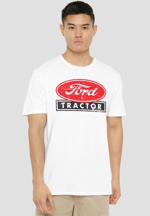 Junger Mann trägt ein weißes T-Shirt mit einem verwitterten roten ovalen Logo mit der Aufschrift "Ford" über einem schwarzen Rechteck mit dem Text "Tractor".