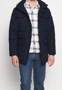 Hombre con chaqueta acolchada azul marino sobre una camisa de cuadros blanca y azul, y jeans azules, de pie contra un fondo liso.