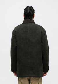 Persona con el cabello trenzado de espaldas, vestido con una chaqueta de cuadros oscuros con cuello de pana y pantalones cargo beiges.