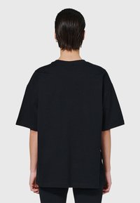 Person med kort vått hår som bär en enkel, oversized svart T-shirt och svarta byxor, sedd bakifrån mot en enfärgad ljus bakgrund.