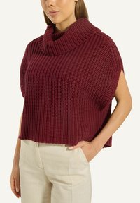 Frau mit einem ärmellosen, dicken, kastanienbraunen Rollkragenpullover und hellbeigen Hosen, mit einer Hand in ihrer Tasche.