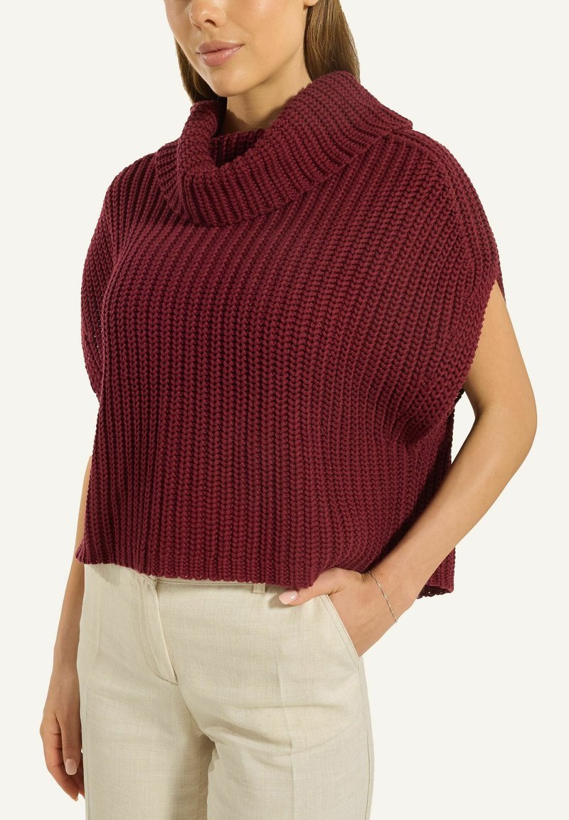 Frau mit einem ärmellosen, dicken, kastanienbraunen Rollkragenpullover und hellbeigen Hosen, mit einer Hand in ihrer Tasche.