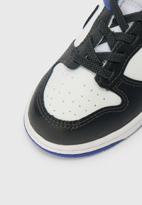 Fekete és fehér sneaker, perforált fehér orral, texturált bőr felsőrésszel, fekete fűzőkkel és lila akcentussal a talpon.
