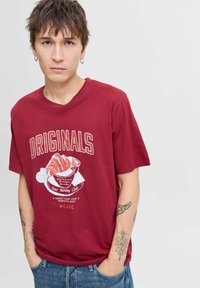 Bordeaux katoenen t-shirt met een grafische print van een croissant en tekst. Korte mouwen, ronde hals, ontspannen pasvorm, met zichtbare tatoeages.