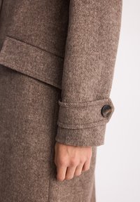 Brązowy wełniany płaszcz o teksturowanym wykończeniu, z detalem mankietu na guziki i smukłą, dopasowaną sylwetką.