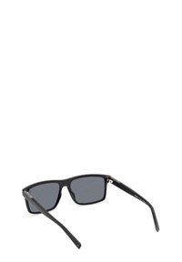 Gafas de sol rectangulares negras con lentes ahumados grises, montura de plástico y acabado brillante. Las varillas presentan acentos metálicos cerca de las bisagras.