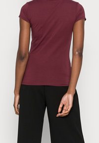 Person iført en tætsiddende burgunder t-shirt med korte ærmer og løse sorte bukser, stående med ryggen til kameraet mod en ensfarvet baggrund.