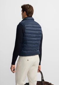 Hombre que lleva un chaleco acolchado azul marino sobre un suéter de punto negro y pantalones beige claros, sosteniendo un bolso de cuero marrón en la mano derecha.