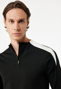 Sort quarter-zip trøje med høj krave og hvide skulderdetaljer, lavet af et glat, strækbart materiale. Strukturert stof omkring halsudskæringen.
