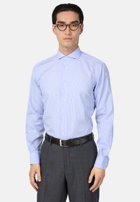 Šviesiai mėlynas dryžuotas marškinėliai su sagčių apykakle, ilgomis rankovėmis ir prigludusia forma, derinami su pilkais kelnėmis ir tamsiu diržu.