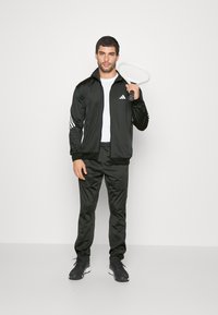 Giacca sportiva nera con strisce bianche, abbinata a pantaloni neri. Il modello tiene una racchetta da tennis e indossa sneaker nere su uno sfondo neutro.