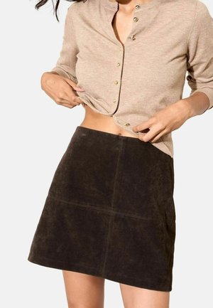 Kobieta ubrana w beżowy kardigan zapinany na guziki, podniesiony do wysokości pępka, oraz ciemnobrązową zamszową minispódniczkę na jednolitym tle.