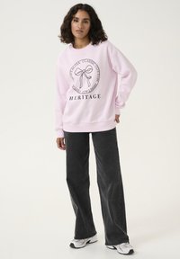 Felpa rosa in cotone, con un grafico circolare che presenta un design a fiocco e la parola "HERITAGE" sotto. Abbinata a jeans scuri.