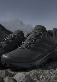 Scarpe da trekking nere da esterno con lacci decorati poggiate su un terreno roccioso con sfondo montano sfocato.