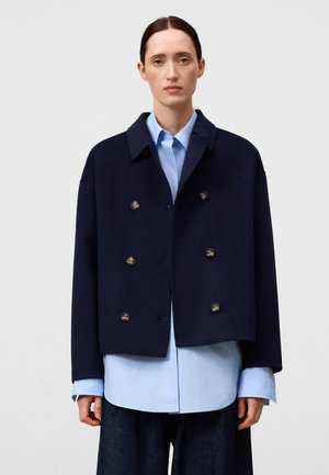 Marineblauwe cropped dubbelzijdige jas met messing knopen, gemaakt van een gestructureerde stof, gelaagd over een lichtblauw shirt met lange mouwen en een kraag.