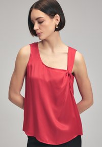 Rød satin top med en enkelt skulderrem, knudedetail ved udskæringen og en flydende silhuet. Glat tekstur og buet kant.