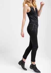 Mono negro con escote en V profundo, cintura ceñida con ojales de metal y piernas ajustadas. Combinado con botas negras hasta el tobillo. Textura suave.