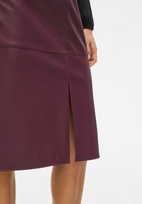 Žena v tmavofialovej koženej sukni po kolená so bočným rozparkom, ktorá odhaľuje časť jej holého stehna a ruku.