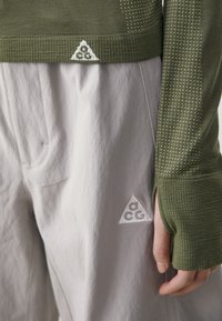 Haut long à manches avec texture en vert olive, featuring des trous pour les pouces, associé à un pantalon gris clair avec un patch logo, tissu lisse et coupe décontractée.