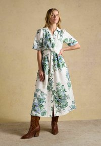 Femeie în picioare cu mâna pe șold, purtând o rochie midi albă cu imprimeu floral verde și albastru și cizme cowboy maro cu toc.