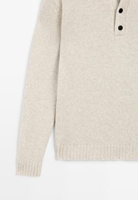 Jasnobeżowy dzianinowy sweter z zaokrąglonym dołem, ściągaczami na mankietach i trzyguzikowym rozcięciem przy dekolcie. Miękka tekstura i minimalistyczny design.