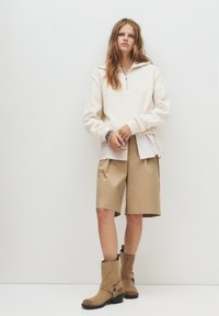 Hellbeige Zip-Up-Fleece-Pullover, der über einem weißen gestreiften Shirt getragen wird, kombiniert mit beigefarbenen knielangen Shorts und braunen Knöchelstiefeln.