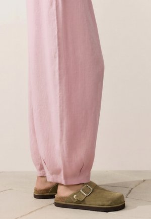 Osoba oblečená do voľných ružových nohavíc a olivovo zelených slip-on nazúvok s prackou stojaca na svetlej dlažbe.
