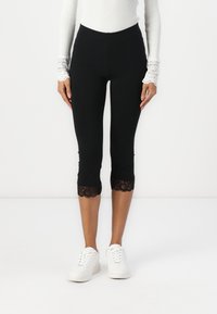 Leggings noires raccourcies avec une bordure en dentelle à l'ourlet, fabriqués à partir d'un matériau extensible. Associés à des baskets blanches et un haut blanc ajusté.