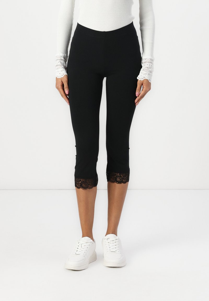Leggings noires raccourcies avec une bordure en dentelle à l'ourlet, fabriqués à partir d'un matériau extensible. Associés à des baskets blanches et un haut blanc ajusté.