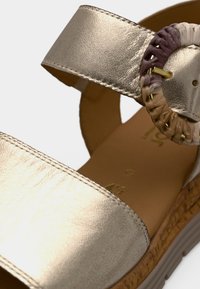 Μεταλλικό χρυσό σανδάλι με δύο φαρδιές λουρίδες και πλεκτή κυκλική διακόσμηση. Υφασμένο πέλμα και ανάγλυφη, ανοιχτόχρωμη εξωτερική σόλα.