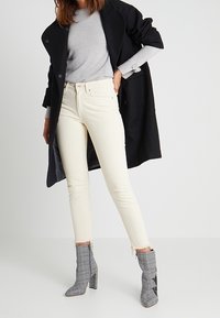 Białe rurkowe dżinsy z surowym wykończeniem, zestawione z szarym swetrem i czarnym płaszczem. Szare kraciaste botki za kostkę z detalami dopełniają stylizację.