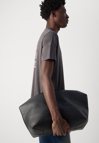 Zwarte leren schoudertas, rechthoekige vorm, gladde textuur, onder de arm gedragen. Gekocht met een grijze T-shirt, zichtbare zij-aanzicht tegen een neutrale achtergrond.