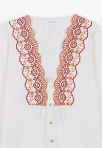 Blouse blanche à col en V profond, ornée d'une broderie florale festonnée bordeaux et orange le long de l'encolure et des boutons sur le devant.