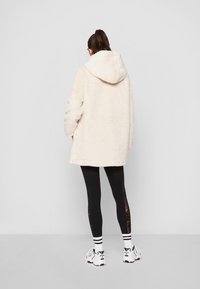 Persona con un abrigo con capucha de piel sintética color crema, leggings negros con paneles de encaje, calcetines blancos con rayas negras y zapatillas blancas, de espaldas.