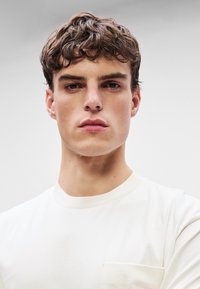 Λευκό βαμβακερό t-shirt με στρογγυλή λαιμόκοψη και τσέπη στο αριστερό μέρος του στήθους. Το μοντέλο έχει κοντά, κυματιστά καφέ μαλλιά και ουδέτερη έκφραση.