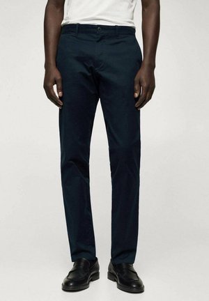 Muž nosící tmavě modré slim fit kalhoty v kombinaci s černými koženými mokasíny a bílou košilí, stojící před jednoduchým pozadím.